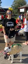 Kate Chapman wearing a shirt that says "YOU HAD ME AT TACOS" and walking a dog that is wearing a taco costume